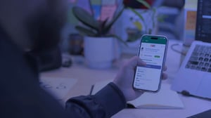Persona sosteniendo un teléfono móvil que muestra una aplicación en pantalla, sobre un escritorio con una laptop, libreta y planta.