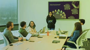 Equipo en reunión colaborativa mientras una persona presenta información en una pantalla con la pregunta ‘¿Qué buscamos?’.