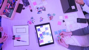 Mesa de trabajo con una tablet, notas adhesivas, maquetas, marcadores y piezas de colores, donde varias personas colaboran en el diseño y planificación de una interfaz digital.