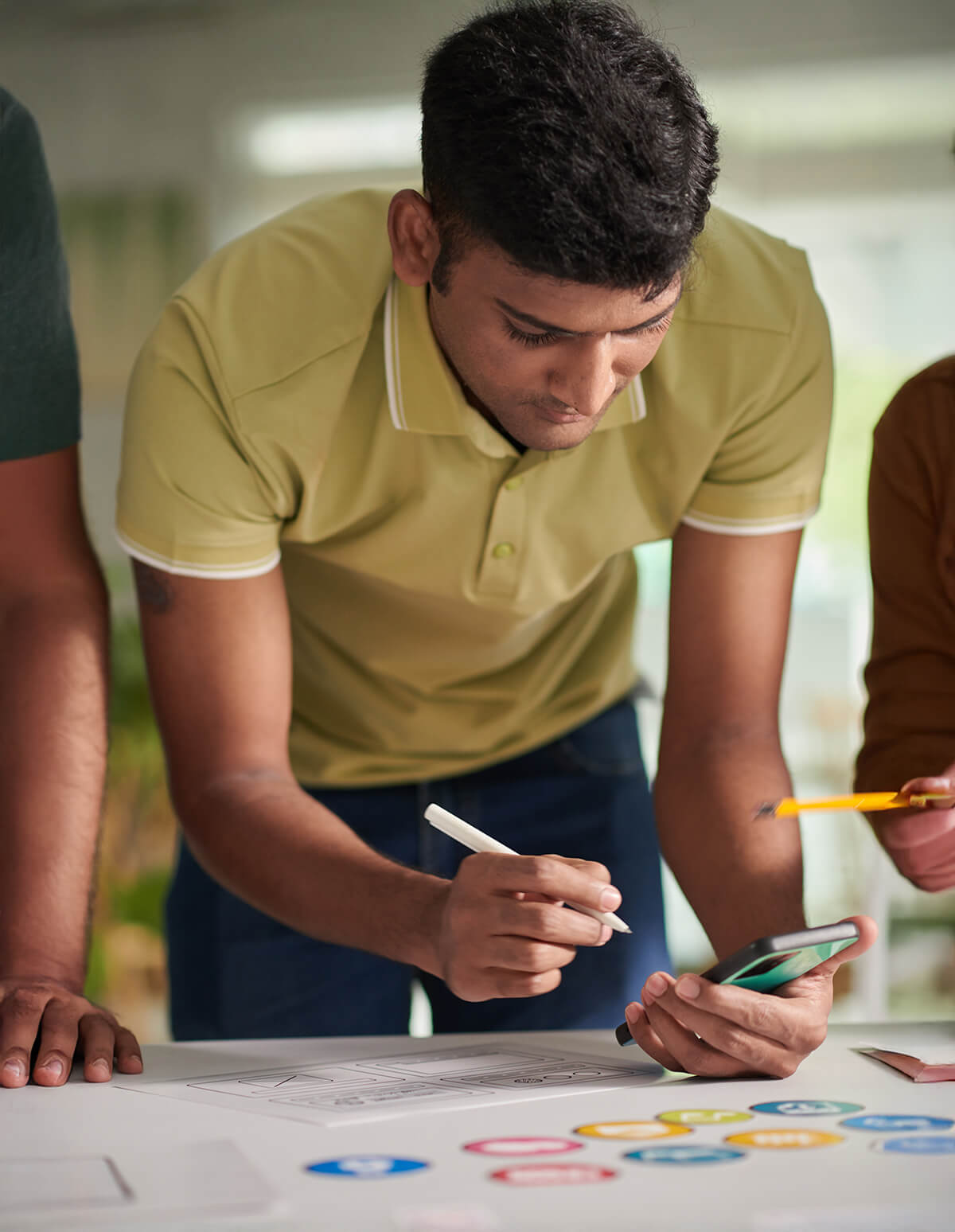 Persona tomando notas y revisando un teléfono móvil mientras colabora en la creación de esquemas y materiales de diseño sobre una mesa.