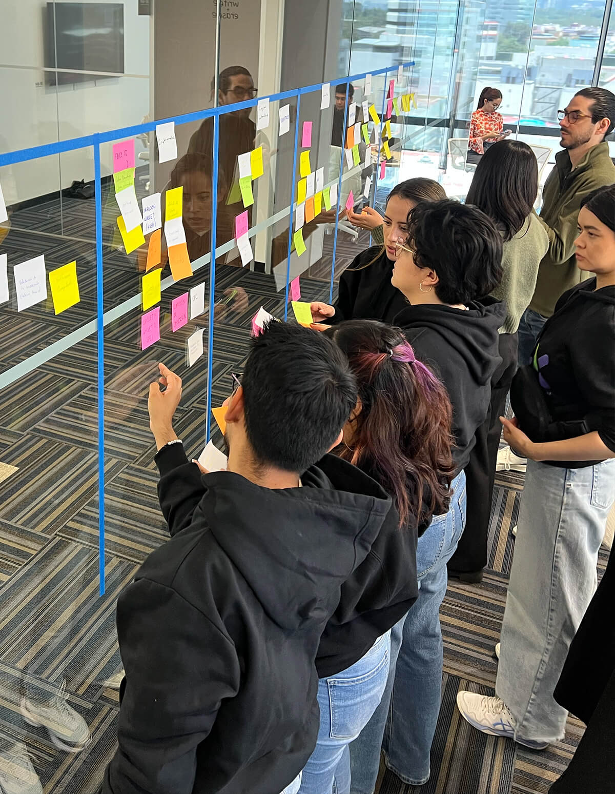 Grupo colaborando frente a un muro de cristal lleno de notas adhesivas, organizando ideas y tareas en una dinámica de trabajo ágil.