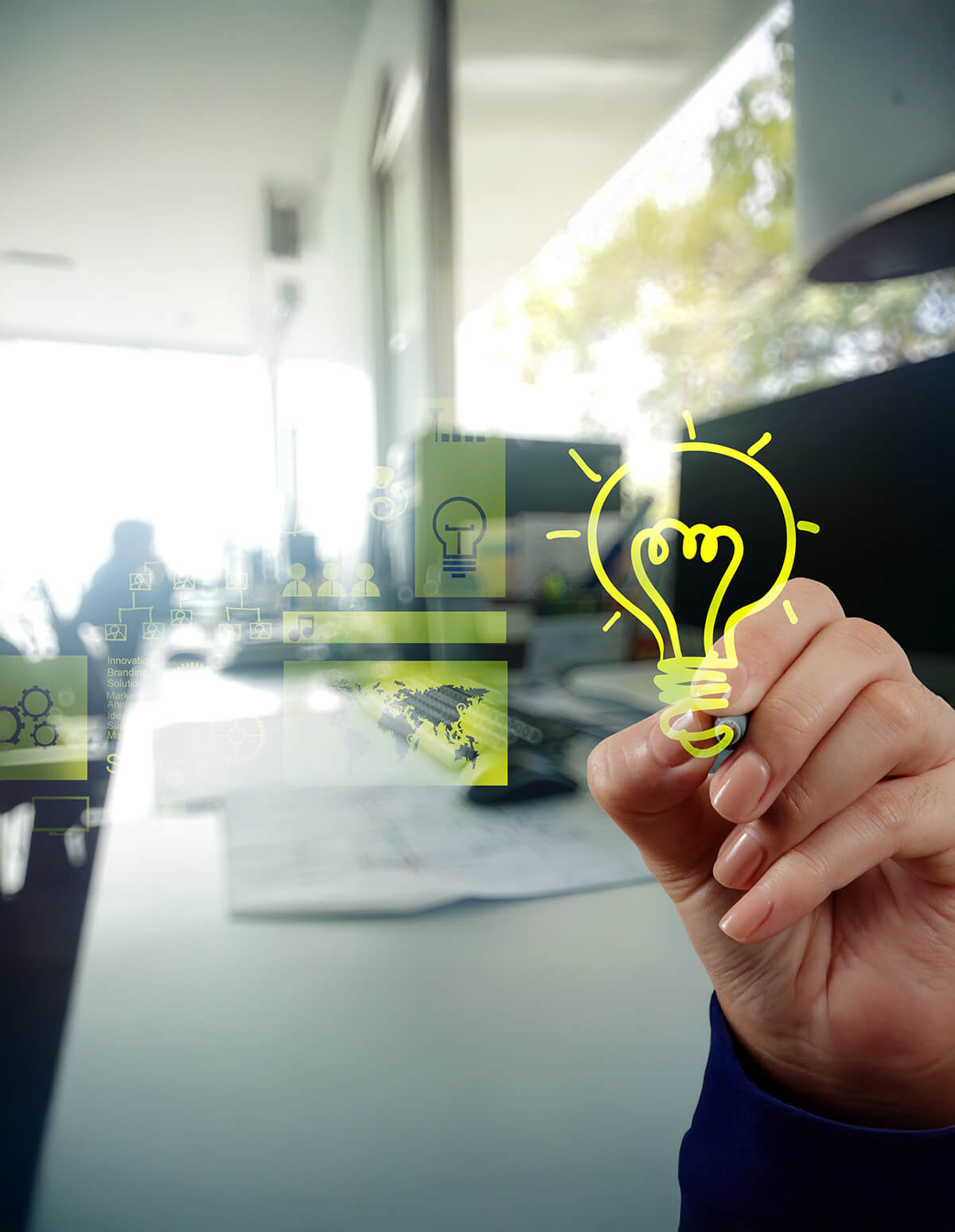 Mano dibujando una bombilla luminosa sobre una superficie transparente, representando ideas e innovación en un entorno de oficina.