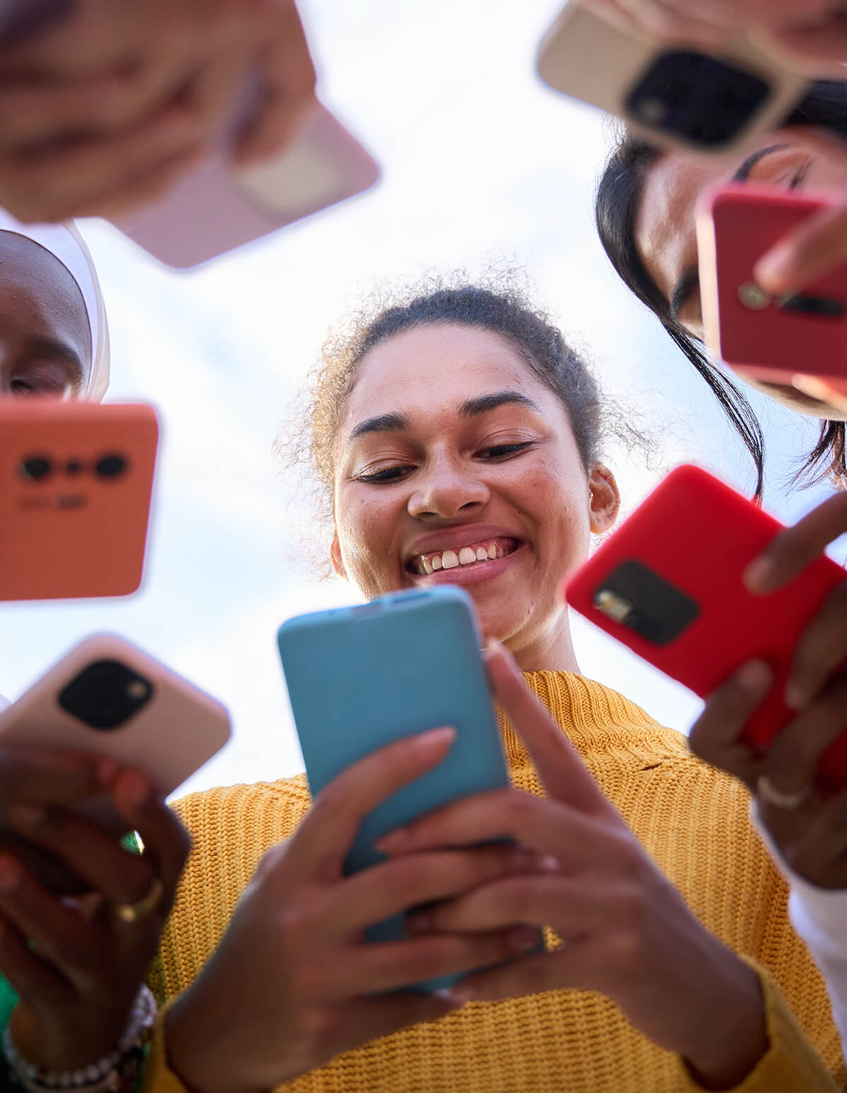 Grupo de personas sosteniendo y usando teléfonos móviles de distintos colores en un círculo, vistos desde un ángulo bajo contra el cielo.